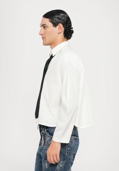 Camisa blanca recortada, de mangas largas, combinada con una corbata negra. Jeans azules con patrones florales bordados. Textura suave, ajuste casual.