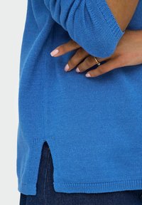 Blå stickad tröja med ribbade muddar och nederkant, försedd med en sidoflik för ökad rörelsefrihet. Bärs över mörka jeans som visar upp texturen.