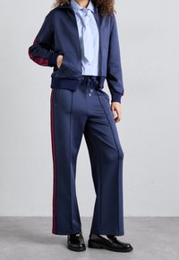 Námořnicky modrá tepláková souprava z hladké látky s červenými proužky na bocích. Obsahuje bundu na zip, volné kalhoty a světle modré tričko pod nimi.