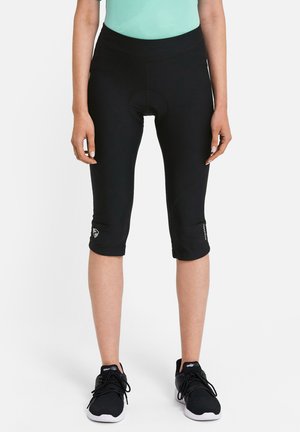 Frau trägt schwarze, knielange Fahrradhosen und schwarze Turnschuhe, steht vor einem schlichten weißen Hintergrund.