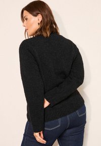 Frau mit braunen Haaren, die goldene Ohrringe, einen schwarzen flauschigen Pullover und dunkelblaue Jeans trägt, vor einem schlichten hellen Hintergrund steht.