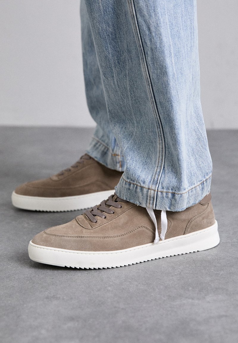 Rjave semiš športne čevlje z belimi gumijastimi podplati, z okroglimi vezalkami in nizkim profilom. Jeani delno prekrivajo čevlje.