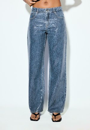 Person iført højbellende, vidde blå jeans med subtilt dyreprint og sorte sandaler med firkantet tå mod en ensartet lys baggrund.