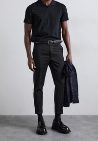 Polo noir, pantalon slim noir avec revers, ceinture noire et chaussures noires brillantes. Tient une veste sombre. Design simple et élégant.