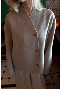 Beige wrap-neule, joka on valmistettu pehmeästä kankaasta, jossa on V-pääntie, pitkät hihat ja neljä pyöreää nappia edessä. Sujuva pinta.