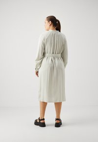 Robe vert clair, longueur genou, avec des rayures verticales, taille ceinturée, manches longues et détail froncé à l'arrière. Associée à des sandales noires.