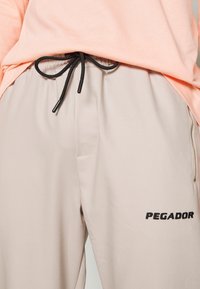 Světlé béžové sportovní kalhoty s černou šňůrkou a boční kapsou na zip. Obsahují vyšívané logo "PEGADOR" v černé barvě. Hladký povrch.