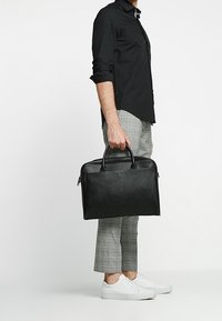 Black leather briefcase with dual top handles, zip closure, and a smooth texture. Features a logo detail on the front.