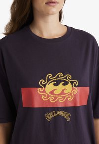 Czarny fioletowy t-shirt z krótkim rękawem, z grafiką żółtej fali i czerwoną prostokątną akcentacją, z napisem "Billabong" w stylizowanym tekście.