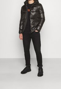 Doudoune noire avec capuche, texture brillante, fermeture éclair et poches avant, portée sur un pull noir à col rond et un jean noir.