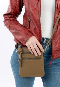 Hnědá kožená crossbody taška s několika zipovými přihrádkami, texturovaným povrchem a odnímatelným popruhem, kterou drží osoba v džínách.