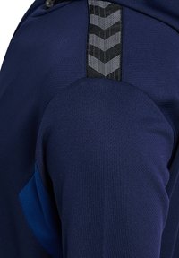 Haut de sport bleu marine en tissu texturé, avec un panneau en chevrons noir près de l'épaule et un accent bleu lisse contrastant.