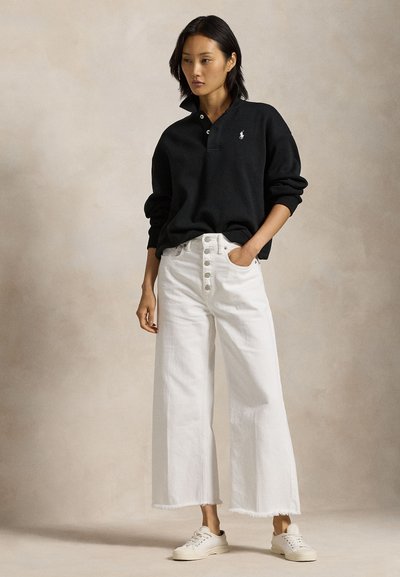 Nő áll, kezét a zsebében, fekete hosszú ujjú pólót, fehér széles szárú, gombos farmert és fehér tornacipőt visel.