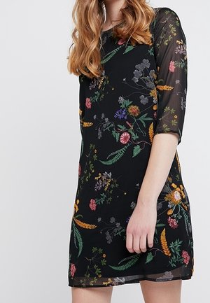 Kvinde iført en sort kjole med gennemsigtige ærmer med farverige blomster- og bregnemønstre, stående mod en ensfarvet lys baggrund.