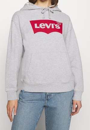 Personne portant un sweat à capuche Levi's gris clair avec un logo rouge et un jean bleu, montré des épaules aux hanches.