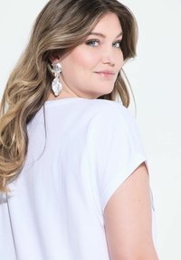 Bílý krátký rukáv top s volným střihem, ukazující zaoblený výstřih, doplněný velkými, lesklými, texturovanými visacími náušnicemi.