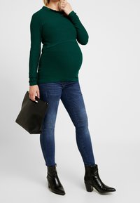 Kobieta w ciąży ubrana w zieloną bluzkę z długim rękawem, niebieskie dżinsy, czarne botki i trzymająca czarną torebkę przy boku.
