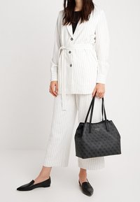 Femme portant un blazer ceinturé blanc à rayures et un pantalon court, tenant un sac fourre-tout Guess à motifs noirs, portant des mocassins noirs.