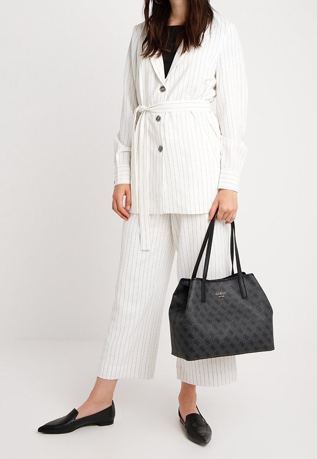 Femme portant un blazer ceinturé blanc à rayures et un pantalon court, tenant un sac fourre-tout Guess à motifs noirs, portant des mocassins noirs.