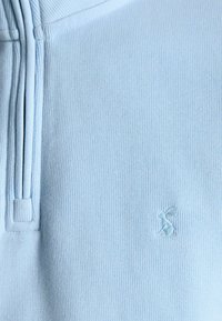 Jasnoniebieska prążkowana tkanina z małym haftowanym logo zająca i częściowo widocznym zamkiem po lewej stronie.