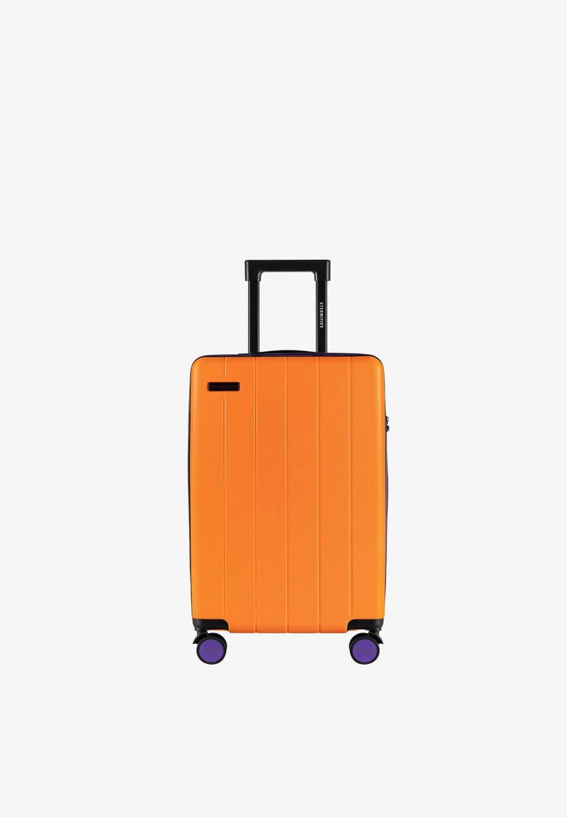 Pomarańczowa walizka z twardą skorupą z pionowymi rowkami, czarna wysuwana rączka i fioletowe kółka. Na przedzie znajduje się małe logo.