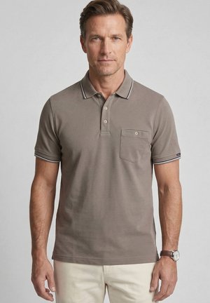 Man wearing brown short-sleeve polo shirt with pocket and beige pants, standing against a plain light background.