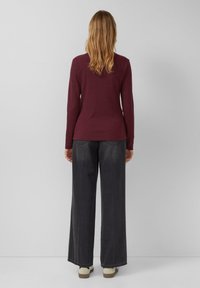 Vrouw die met haar rug naar de camera staat, gekleed in een bordeauxrode langmouwtop, brede donkergrijze jeans en lichtgekleurde schoenen tegen een effen achtergrond.