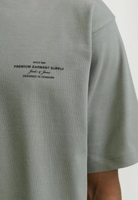 Siva bombažna majica s kratkimi rokavi, s temnim potiskom logotipa, ki vključuje ime blagovne znamke in oblikovalske podrobnosti, gladka tekstura, sproščen kroj.