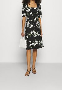 Kobieta w czarnej, kwiecistej sukience do kolan z rękawami do łokci, trzymająca małą białą torebkę i nosząca czarne sandały na obcasie.