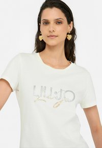 T-shirt blanc à manches courtes avec des lettres texturées en paillettes argentées et dorées. Encolure ronde, en coton doux et coupe décontractée.