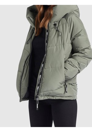 Vrouw draagt een olijfgroene gewatteerde jas met een hoge kraag, ritssluiting, zijzakken en zwarte voering, gecombineerd met een zwarte broek.