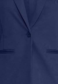 Blazer azul marino con una textura suave, cierre de un solo botón, dos bolsillos frontales y detalles de diseño a medida.