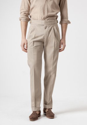 Hombre con pantalones beige de cintura alta y plisados, con bajos doblados, combinados con sandalias marrones de ante trenzado y una camisa beige remangada.