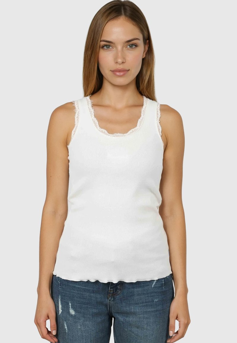 Femme aux cheveux bruns clairs et raides, portant un haut blanc sans manches avec des bordures en dentelle et un jean bleu foncé déchiré, faisant face à l'avant.