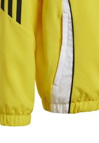 Giacca atletica gialla con polsini elasticizzati, caratterizzata da un design a tre strisce nere e una tasca con zip bianca. Texture liscia e materiale leggero.