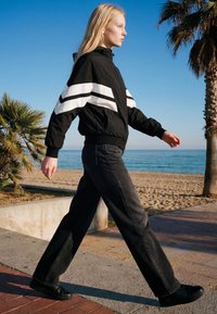 Noor blondide pikkade juustega naine jalutab rannarajal, kandes musta jope valgete triipudega ja musti pükse selge taeva all.