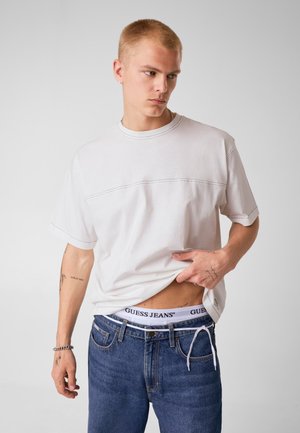 Junger Mann mit kurzen blonden Haaren trägt ein weißes T-Shirt und blaue Jeans, hebt das Shirt an, um den weißen Bund von Guess Jeans zu zeigen. Neutraler Hintergrund.