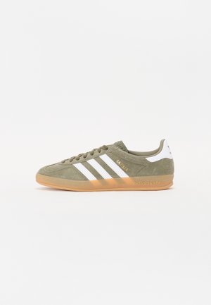 Baskets Adidas Gazelle vert olive avec des bandes blanches, semelle en gomme et languette de talon blanche sur un fond blanc uni.