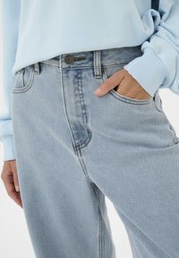 Světle modré džínové kalhoty s rukou v přední kapse, doplněné o sladěný světle modrý dlouhý rukáv.