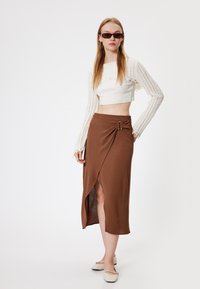 Bruine wikkelrok met zijaccenten en een hoge split, gecombineerd met een witte cropped gebreide top en crèmekleurige platte schoenen. Aanvoelt glad en gemaakt van lichtgewicht stof.
