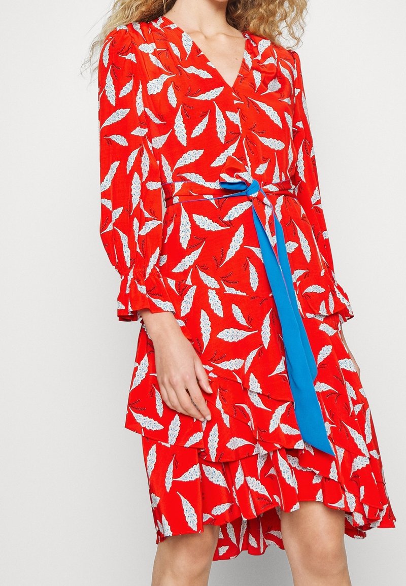 Rotes Kleid mit weißen Blattmustern, langen Puffärmel, tiefem V-Ausschnitt und einem blauen Stoffgürtel, der in der Taille gebunden ist. Rüschensaum-Detail.