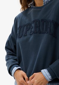 Persona vistiendo un sudadera azul marino de Superdry sobre una camisa de botones a rayas azules y blancas, con las manos entrelazadas a la altura de la cintura.