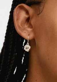 Boucle d'oreille en argent avec une grande pierre précieuse facettée et transparente, encastrée dans un boîtier en métal décoratif, suspendue près d'une coiffure tressée.
