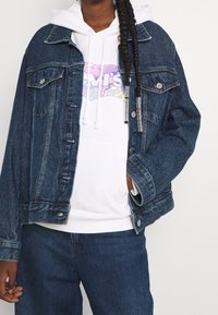 Persona con trenzas que lleva una sudadera blanca con un logo colorido debajo de una chaqueta de mezclilla azul oscuro y jeans azules, con una mano en el bolsillo.