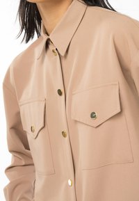 Chemise beige à boutons avec deux poches poitrine, chacune sécurisée par un seul bouton pression doré, portée par une personne aux cheveux foncés.