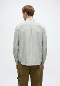 Ανοιχτό πράσινο ριγέ πουκάμισο με μακριά μανίκια και γιακά, συνδυασμένο με χακί παντελόνι καμπαρντίνα. Ομοιόμορφες κατακόρυφες ρίγες με μανσέτες κουμπωτές.