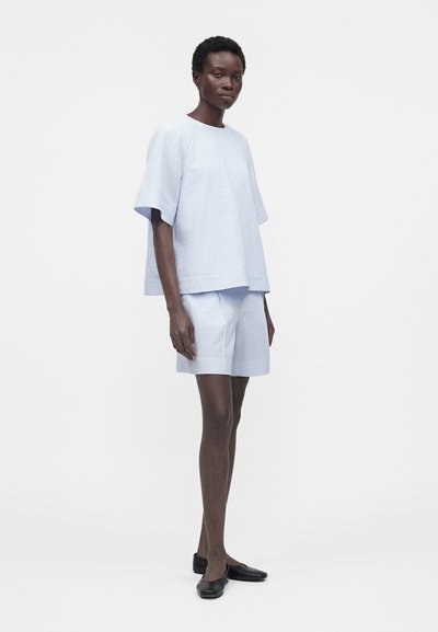 Γυναίκα όρθια, φοράει ανοιχτό μπλε υφασμάτινη μπλούζα με κοντά μανίκια και ταιριαστό σορτς με μαύρα επίπεδα παπούτσια μπροστά σε λευκό φόντο.