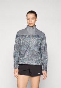 Kurtka typu windbreaker z szaro-niebieskim wzorem w kamuflaż, z zamkiem z przodu, stójką i długimi rękawami; w zestawie z czarnymi szortami.