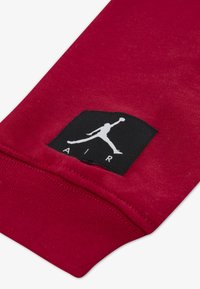 Κοντινό πλάνο μανσετών κόκκινου μανικιού με μαύρο ορθογώνιο λογότυπο που δείχνει το λευκό λογότυπο Jumpman και τη λέξη «AIR» από κάτω.