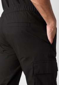 Schwarze Cargo-Hosen mit einem elastischen Bund, Seitentaschen und flacher Gesäßtasche. Der Stoff hat eine glatte Textur und ein strapazierfähiges Design.